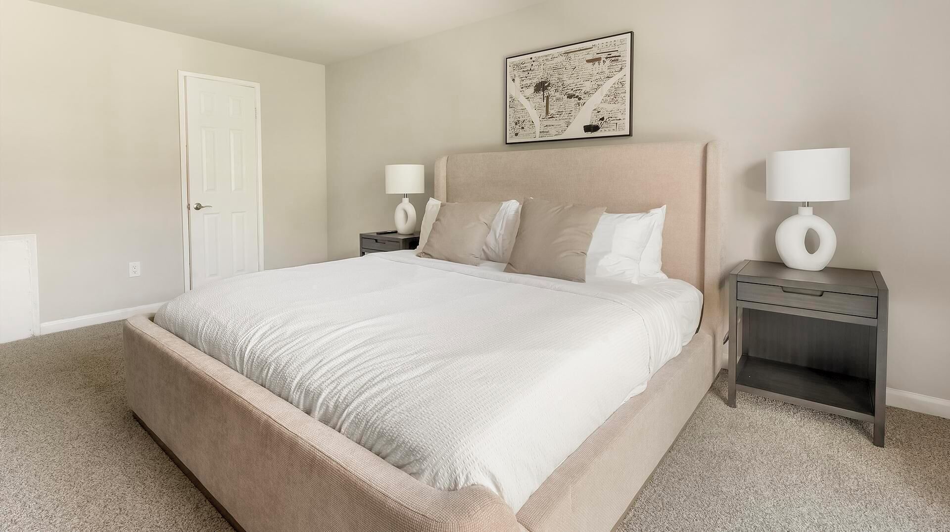 A neatly arranged bedroom with a large bed, two bedside tables with lamps, a door, and a framed artwork on the wall.
