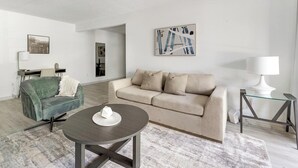 A modern living room with a green armchair, a beige sofa, a glass coffee table, and a white lamp on a glass table.
