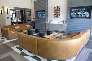 Ein modernes Wohnzimmer mit einer großen, winkelförmigen Couch, einem Couchtisch und Poufs. Zwei Flachbildfernseher sind an der Wand montiert, und im Hintergrund ist eine Küchenzeile zu sehen.