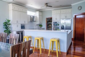 Une cuisine moderne avec des meubles blancs, un îlot central doté de tabourets, des appareils en acier inoxydable et un ventilateur de plafond.