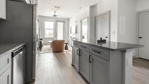 A modern kitchen with grey cabinets, a built-in oven, and a large island. There is a dining area with a round table and chairs, and a large window providing natural light.