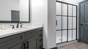 Ein modernes Badezimmer mit weißer Ablagefläche, zwei Waschbecken, einem großen Spiegel und einer Duschkabine.