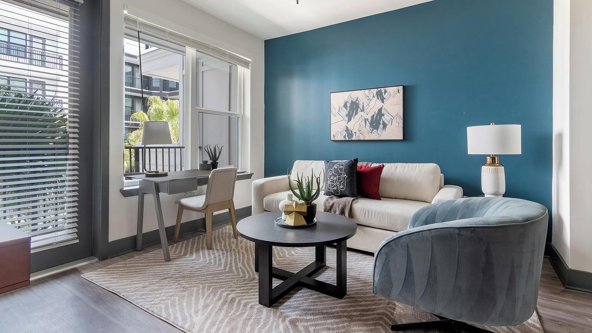A modern living room with a blue accent wall, a sofa, a chair, a coffee table, and a desk with a lamp.