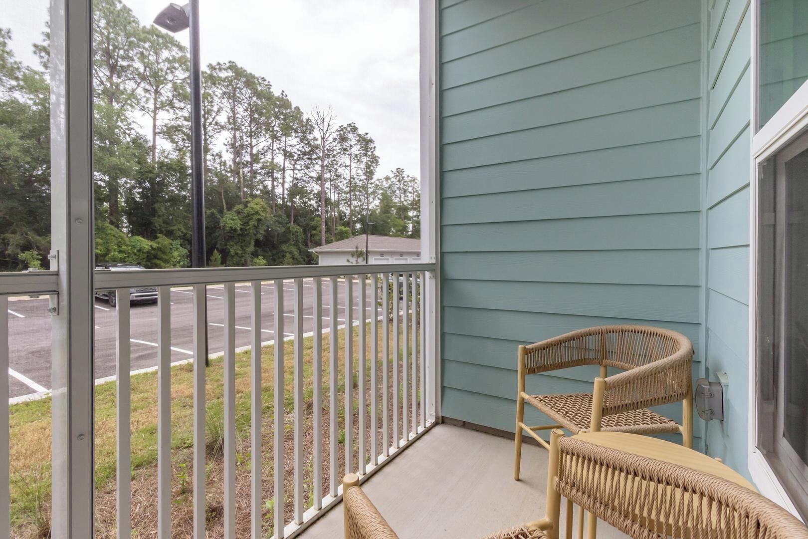 A balcony with a wicker chair and a small table, overlooking a parking lot and trees.