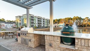 An outdoor kitchen area with a grill, a water feature, and a view of a multi-story building in the background. - Landing | Incredible 1BD, Gym, Yoga Studio (Santa Rosa Beach)