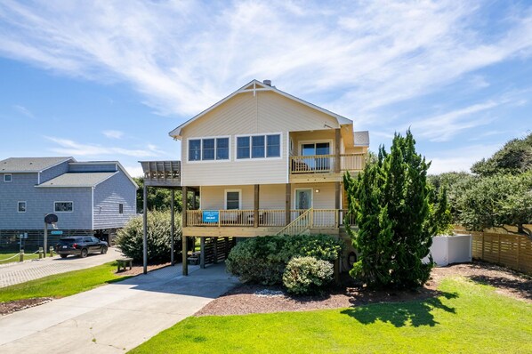 A two-story building with a balcony on the upper floor, a covered outdoor area, and a driveway leading to a parked car.
