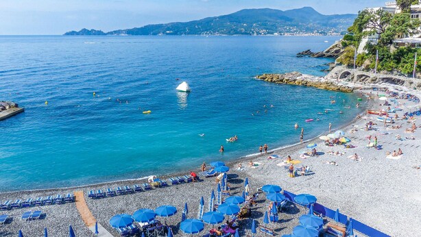A beach with clear blue waters, a rocky shoreline, and a view of distant hills.