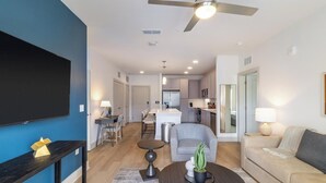 A modern living room with a flat-screen TV, a ceiling fan, a dining area with a table and chairs, a kitchen with a refrigerator and microwave, and a sofa with a lamp. - Landing | Spectacular 2BD, Pool, Yoga Studio (Santa Rosa Beach)