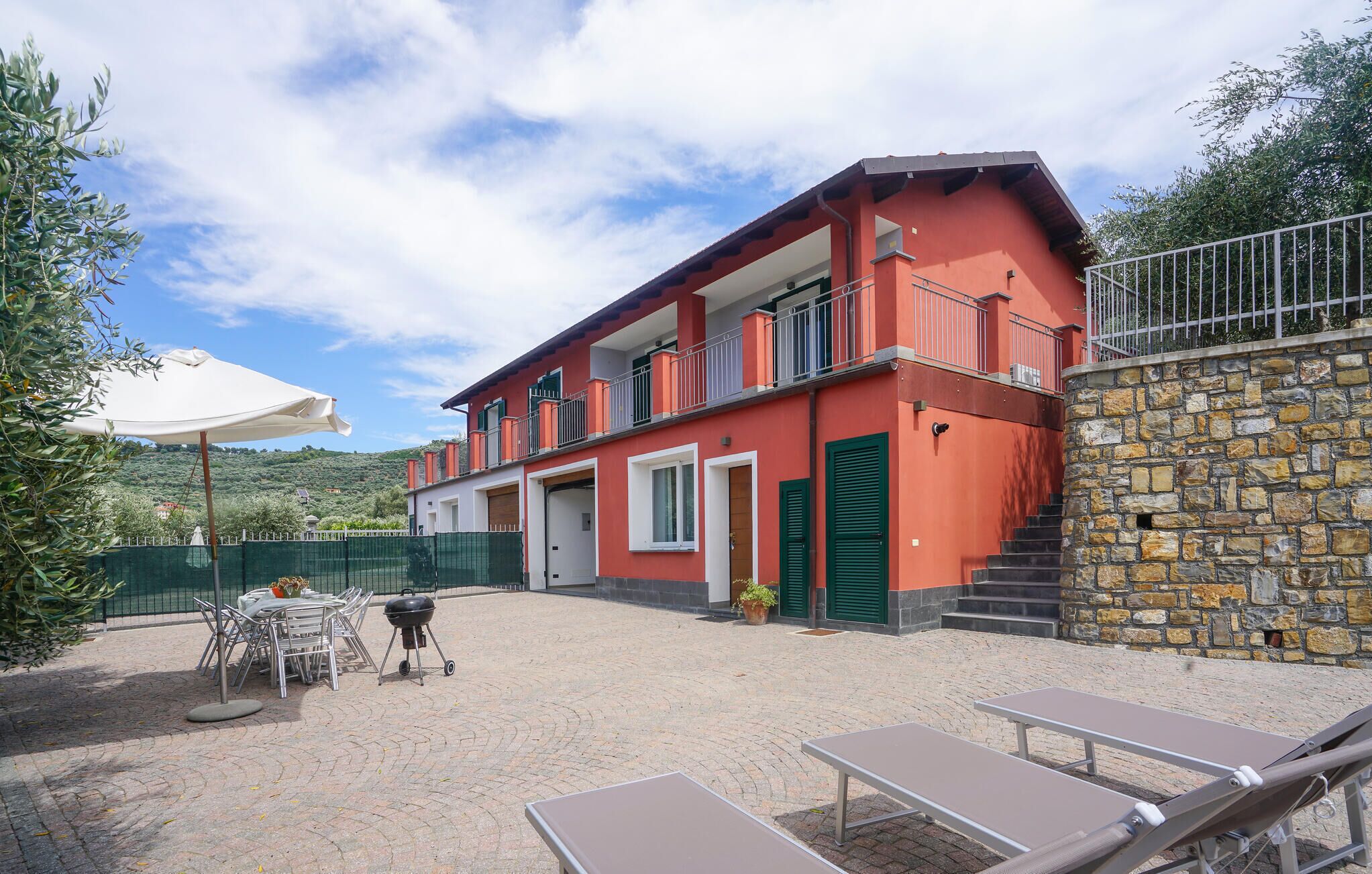 A two-story building with a red facade, a stone wall, outdoor seating, and a barbecue grill.