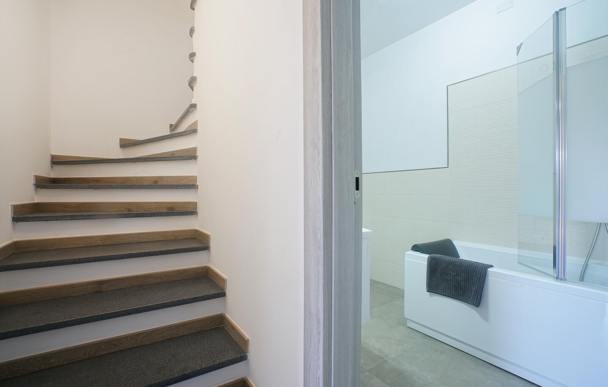 Una escalera con escalones de madera y un vidrio que da a un baño con una bañera blanca y una toalla colgada en la pared.