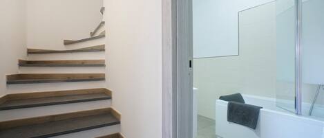 Una escalera con escalones de madera y un vidrio que da a un baño con una bañera blanca y una toalla colgada en la pared.
