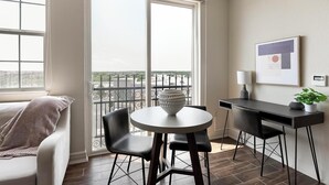 A modern room with a balcony, a round table with chairs, a desk with a lamp, and a framed picture on the wall.