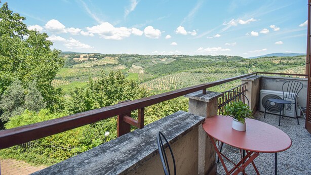 Un balcon avec une table rouge, un pot de fleurs et une chaise. Le balcon offre une vue sur un paysage verdoyant, avec des collines ondoyantes et un ciel dégagé.