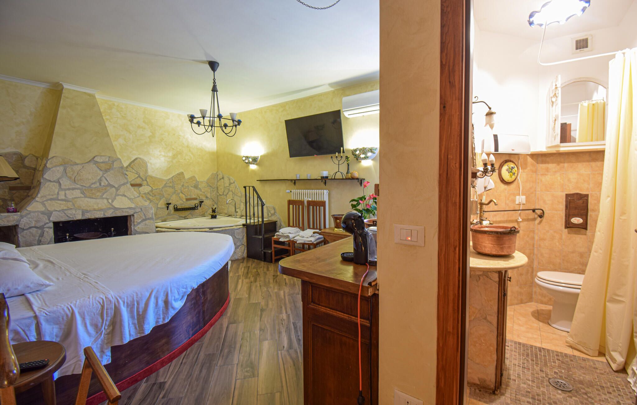 Une chambre avec une cheminée en pierre, un grand lit, un lustre et un téléviseur fixé au mur.
