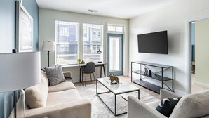 A modern living room with a sofa, coffee table, TV, and a desk with a chair. - Landing | Stylish 2BD, Pool, Gym (Leland)