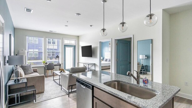 A modern kitchen with a granite countertop, stainless steel sink, and overhead lighting. - Landing | Stylish 2BD, Pool, Gym (Leland)