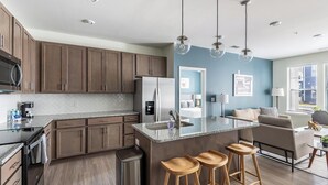 A modern kitchen with dark wood cabinets, a stainless steel refrigerator, and a granite countertop. There are three wooden stools at the island, and the room has a blue accent wall. - Landing | Stylish 2BD, Pool, Gym (Leland)