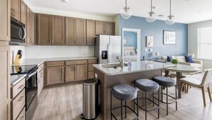 A modern kitchen with wooden cabinets, stainless steel appliances, a granite countertop, and a bar stool area. - Landing | Spectacular 2BD, Pool, Gym (Leland)