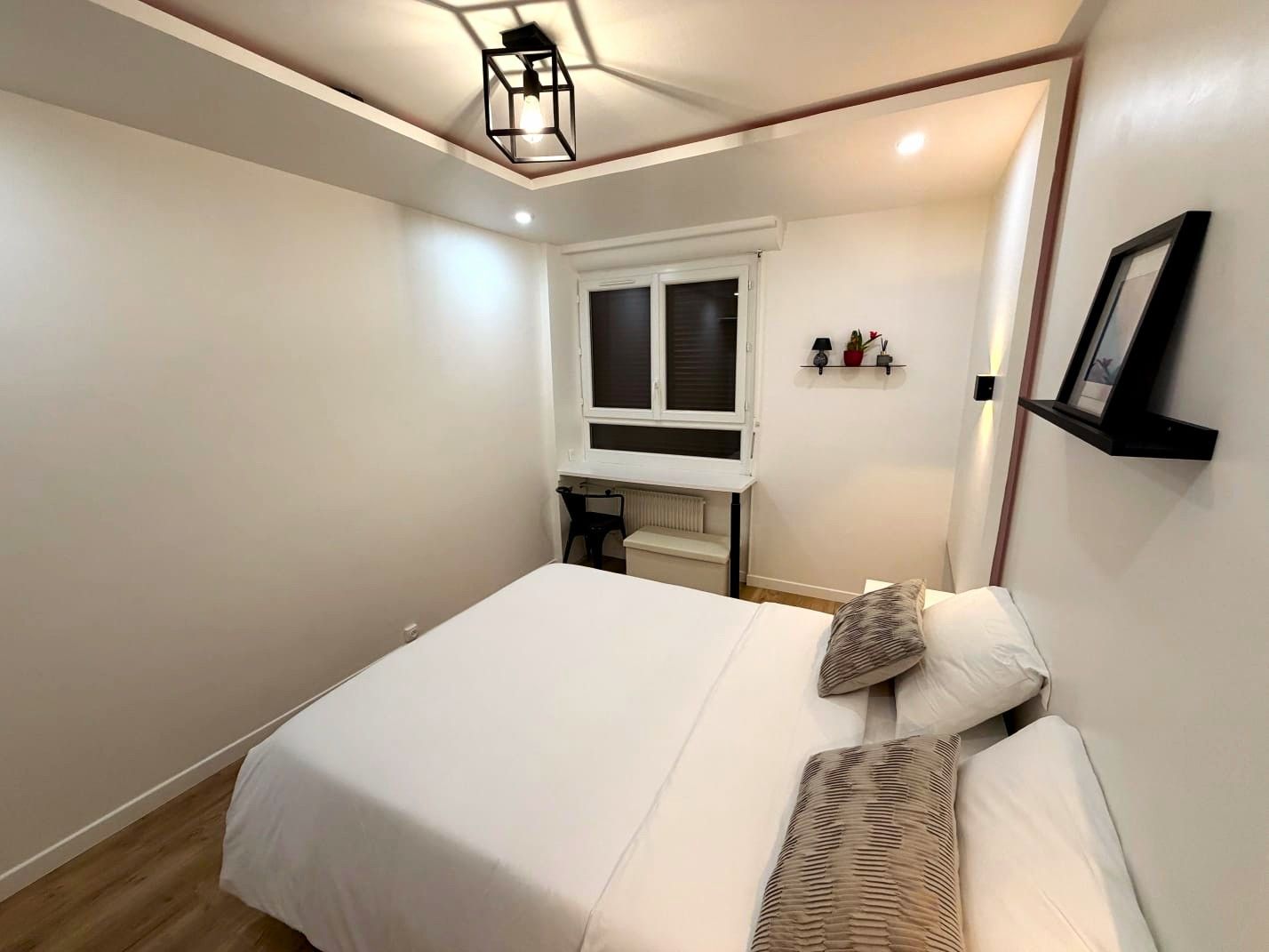 A bedroom with a bed, a window, a radiator, a shelf with decorative items, and a wall-mounted picture frame.