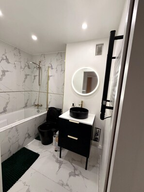 Une salle de bain avec une baignoire, un grand miroir rond avec éclairage intégré, un lavabo noir et un meuble-lavabo noir.