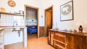 A kitchen with a sink, stove, and wooden cabinets. There is a bed with a blue blanket and a wall clock.