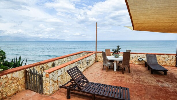 A terrace with a stone wall, a wooden lounge chair, a table with chairs, and a view of the sea.