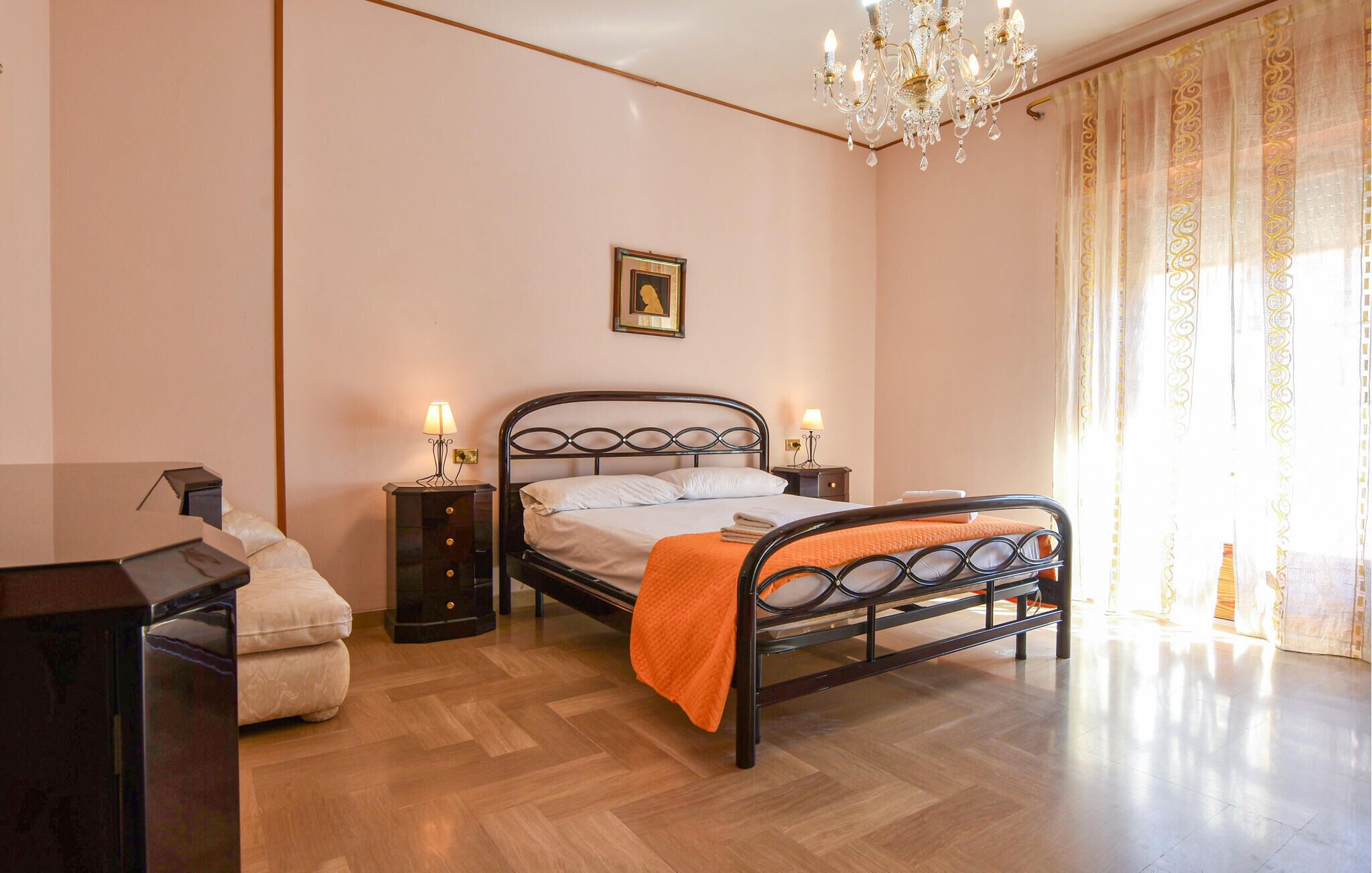 A bedroom with a bed, bedside tables, a desk, a chair, a chandelier, and a window with curtains.