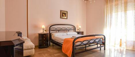 A bedroom with a bed, bedside tables, a desk, a chair, a chandelier, and a window with curtains.