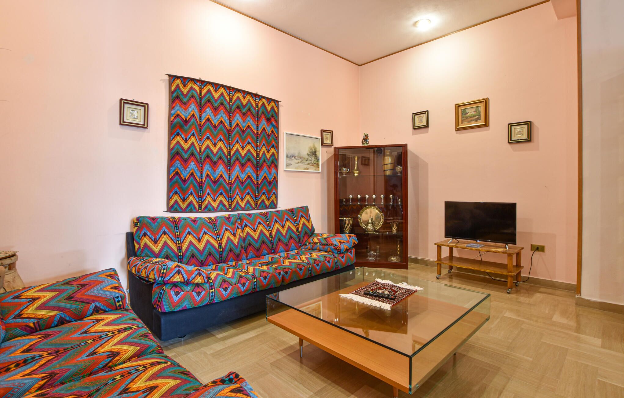 Ein Wohnzimmer mit einem farbenfrohen, zickzack-gemusterten Sofa, einem Glastisch, einem Fernseher auf einem Holzschrank und einer Vitrine.
