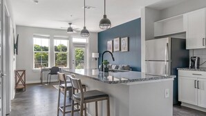 A modern kitchen with white cabinets, a large island with bar stools, and a view of the outdoors through large windows.