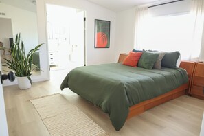 Ein modernes Schlafzimmer mit einer grünen Tagesdecke, einem Holznachttisch, einer Topfpflanze und einem großen Fenster mit Vorhängen.