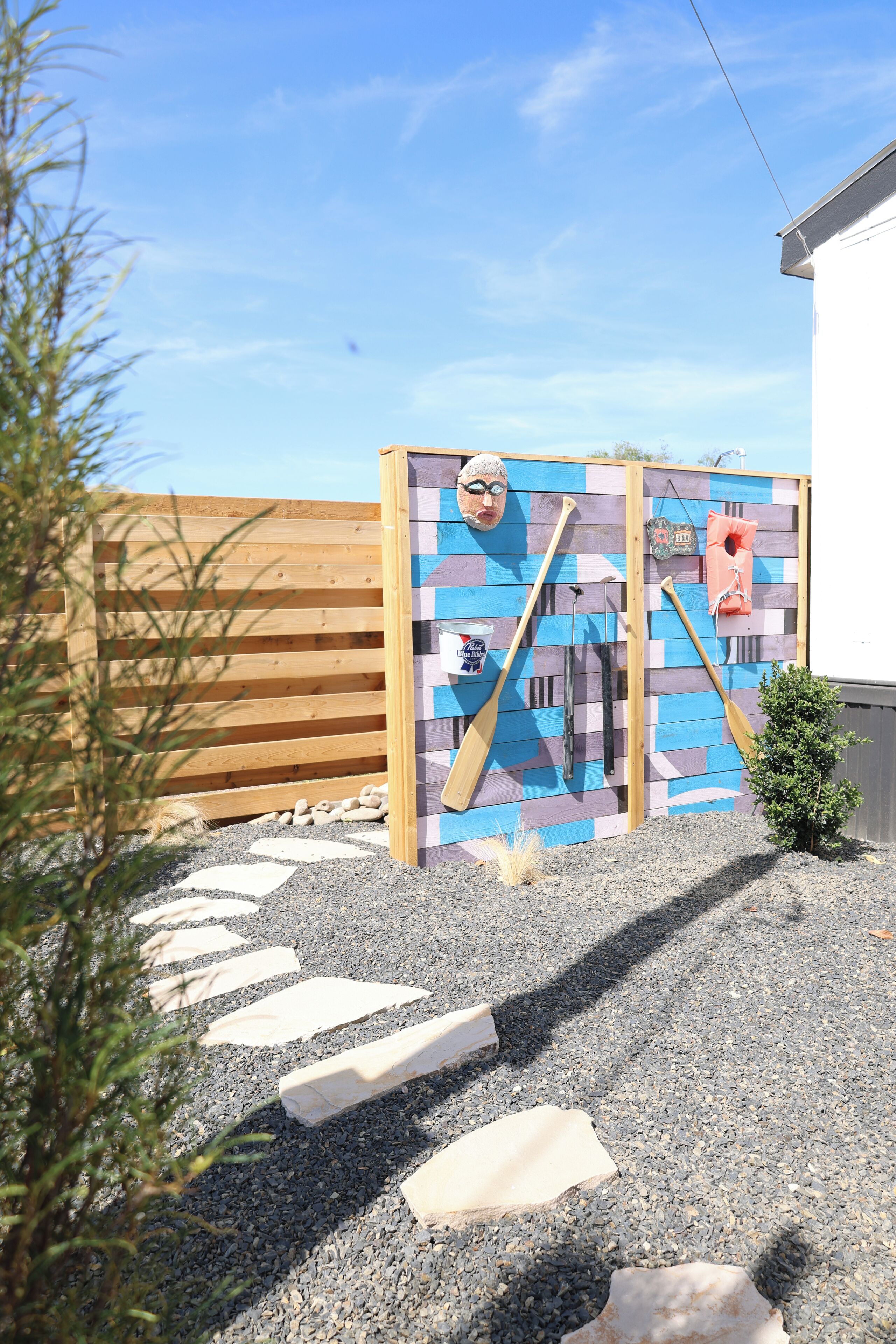 Une clôture en bois, un mur coloré orné de divers objets et un petit arbuste.