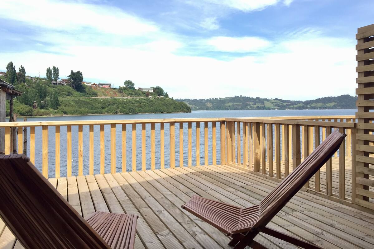 A wooden deck with two lounge chairs facing a calm lake, surrounded by a wooden railing.