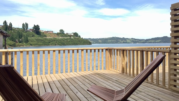 A wooden deck with two lounge chairs facing a calm lake, surrounded by a wooden railing. - Palafito Azul Apart Hotel (Castro)
