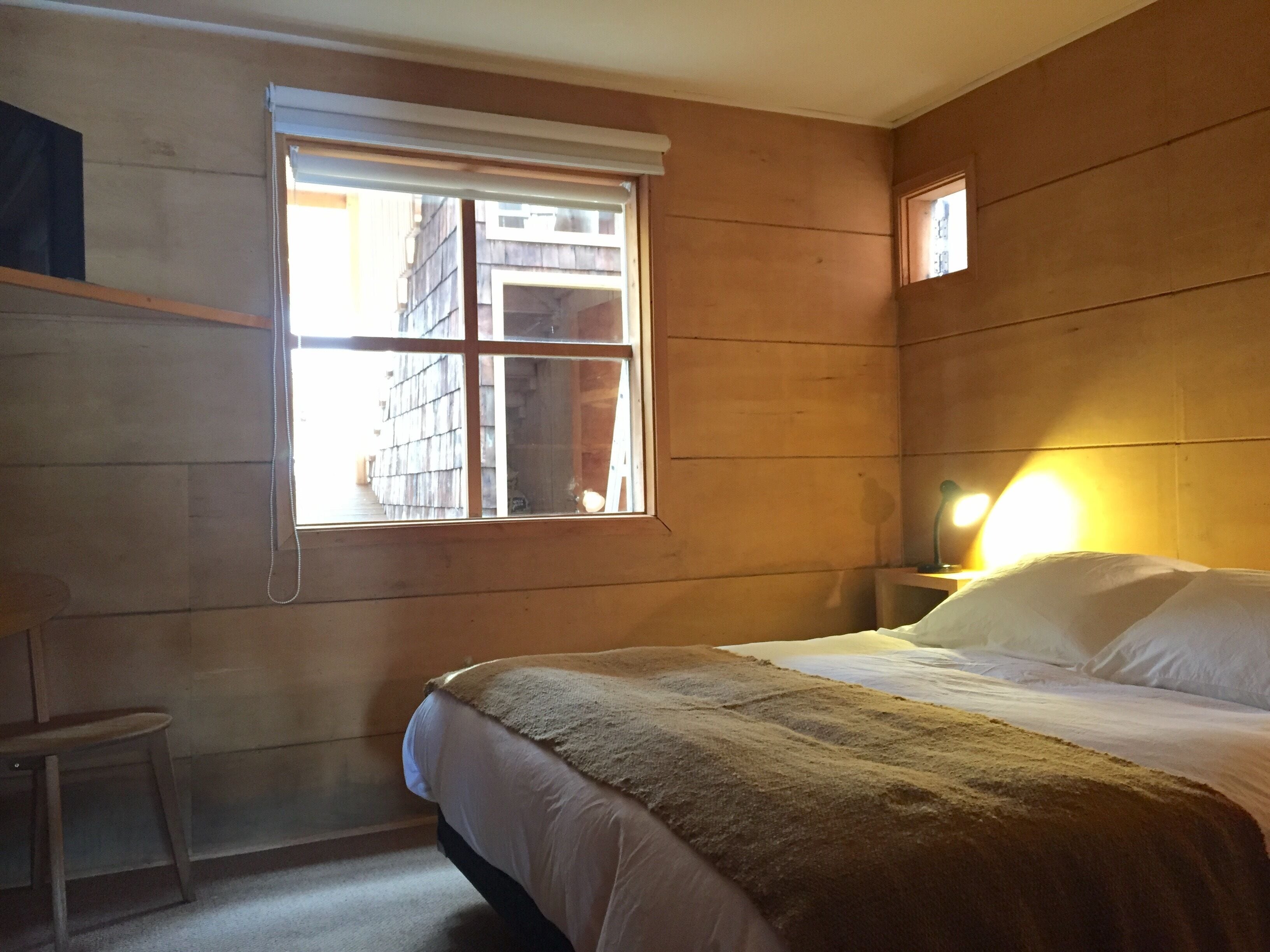 Una habitación con cama, una mesita, una ventana y una mesita de noche con lámpara.