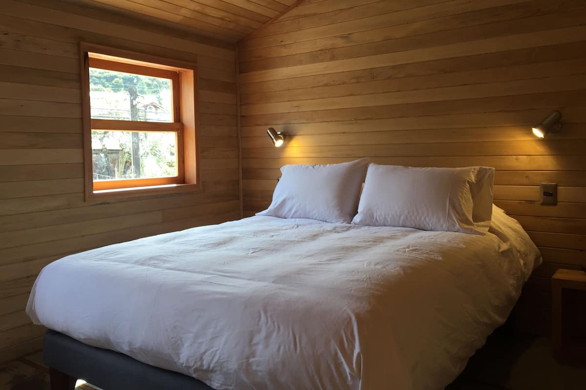 A bedroom with a wooden ceiling, a bed with white bedding, a window with a view of greenery, and wall-mounted lighting.