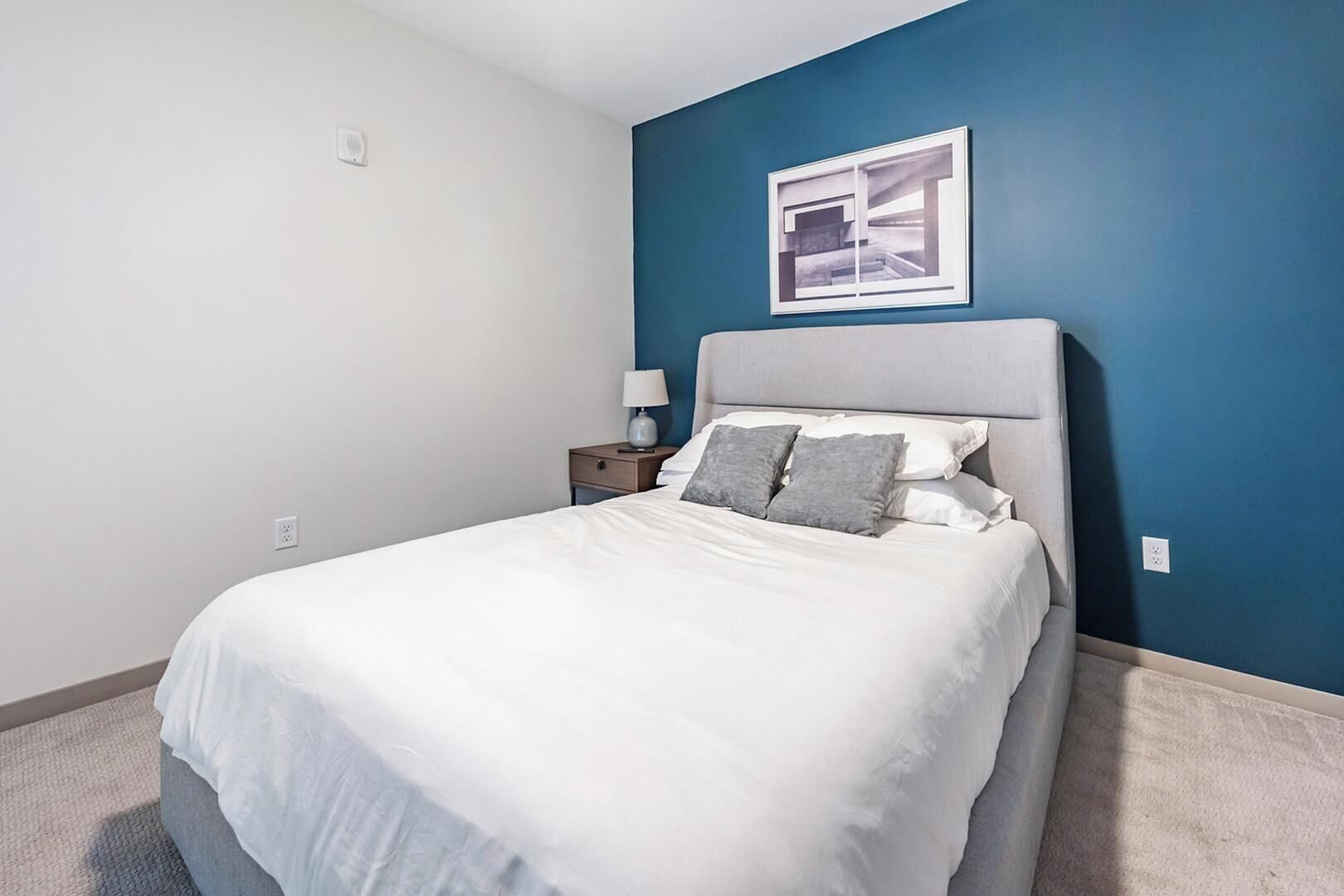 Quarto com cama, mesa de cabeceira, abajur e um quadro na parede.
