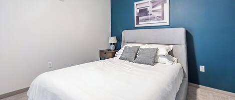 Quarto com cama, mesa de cabeceira, abajur e um quadro na parede.