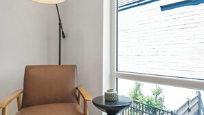 A room with a large window, a brown chair, a small table with a decorative item, and a floor lamp.