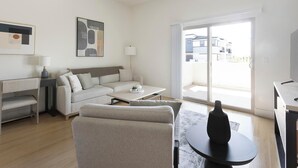 A modern living room with a sofa, a coffee table, a side table, and a lamp. There is a large window with a view of buildings outside. - Landing | Incredible 2BD, Pool (Casas Adobes)