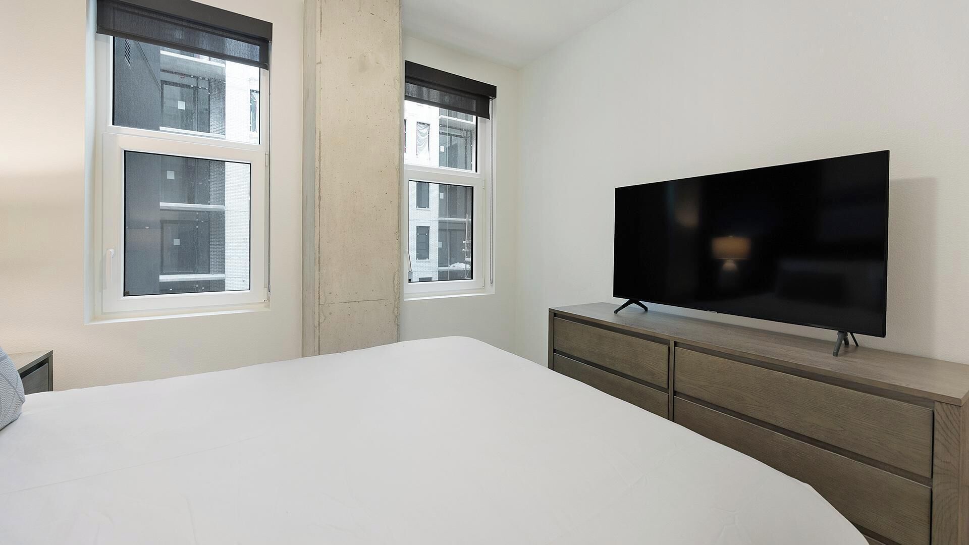 Une chambre à coucher avec un grand lit, un téléviseur posé sur une commode en bois et deux fenêtres équipées de stores.