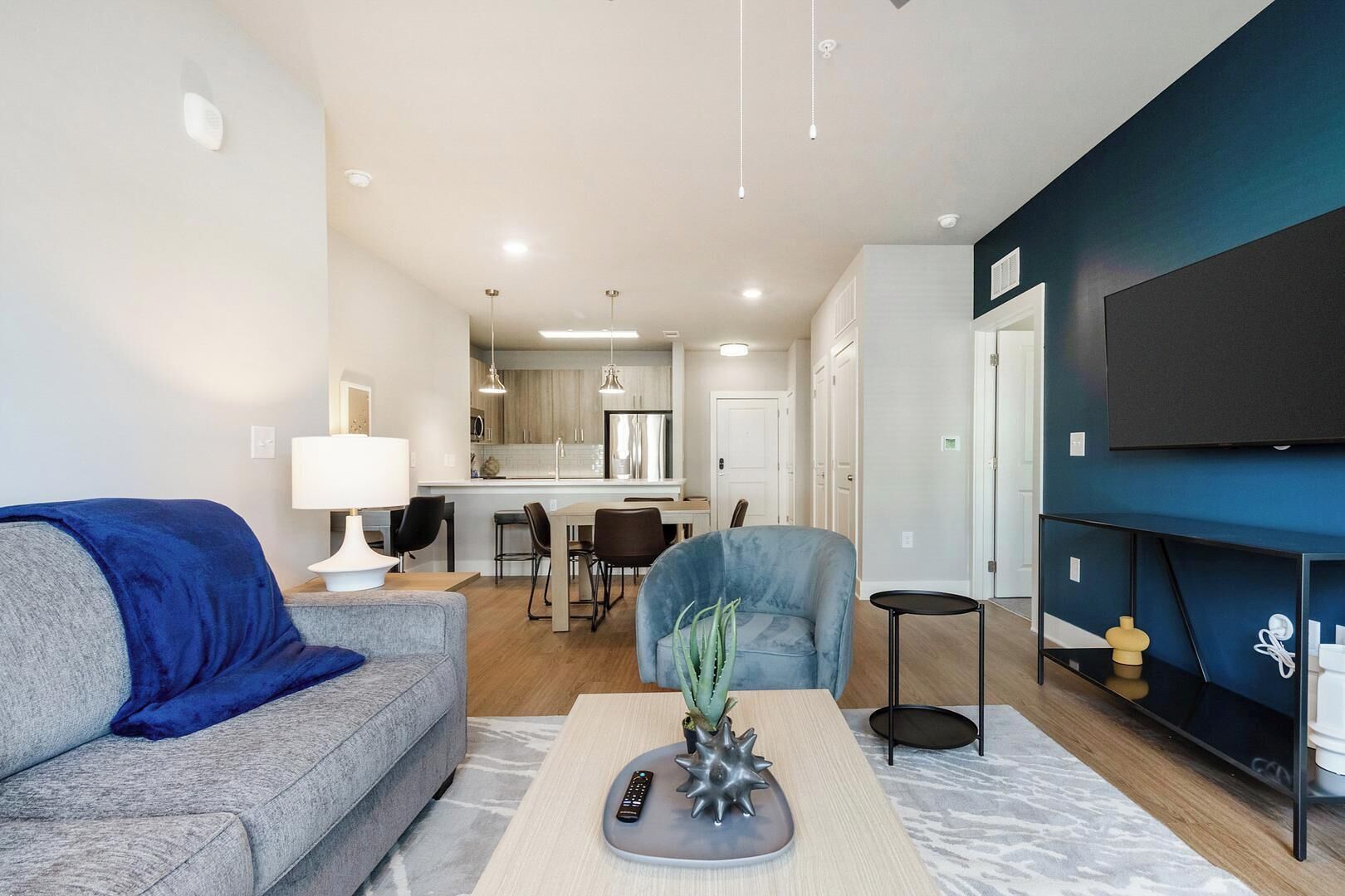 A modern living room with a grey sofa, a blue armchair, a coffee table with a remote control, and a TV mounted on a dark blue wall.