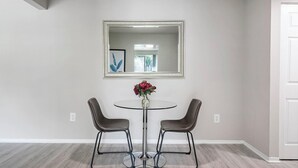 Een eetruimte met een ronde glazen tafel, twee stoelen, een vaas met bloemen en een grote spiegel aan de muur.