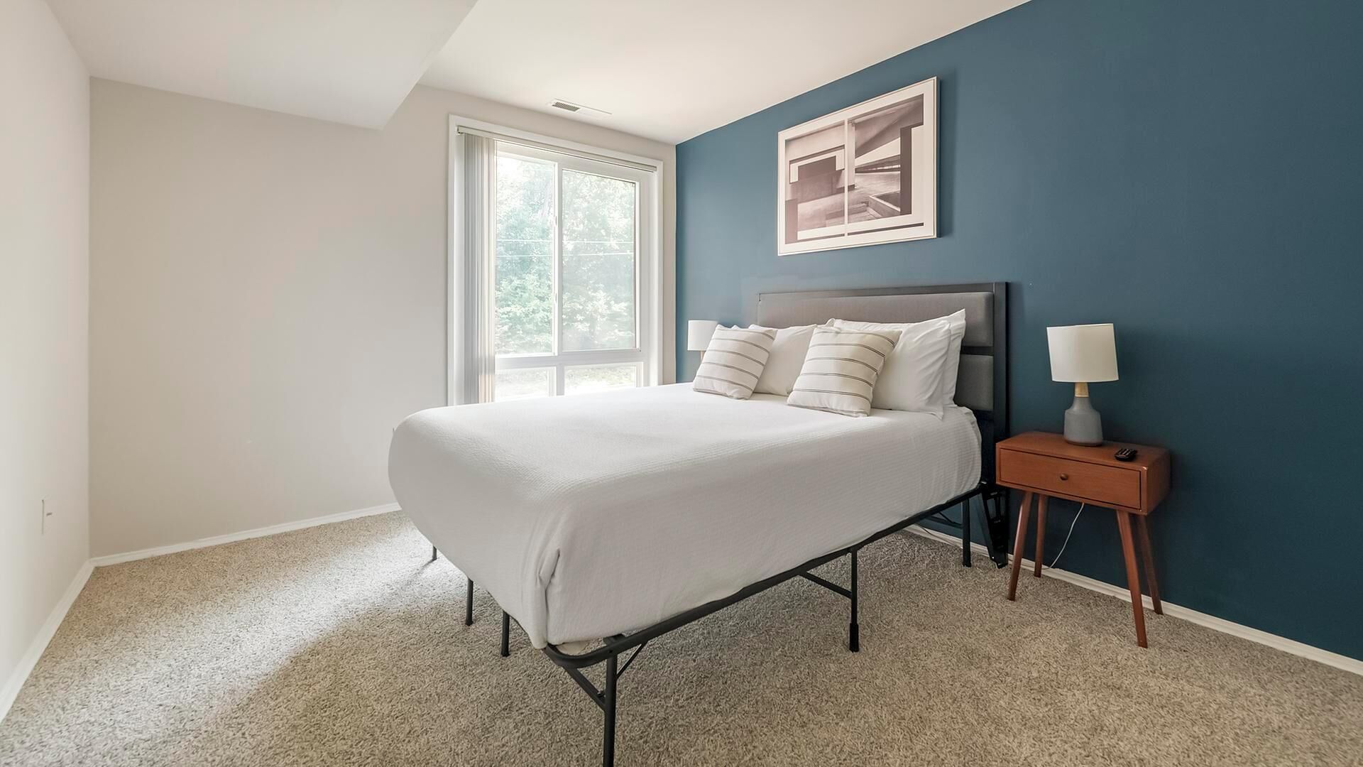 A bedroom with a bed, a nightstand, a lamp, a window, and a framed picture on the wall.