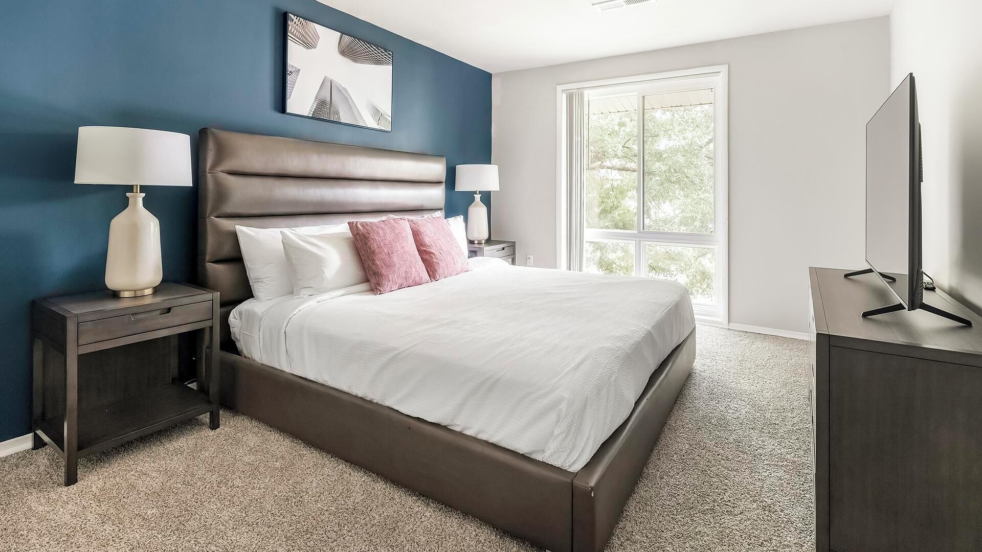 A modern bedroom with a large bed, two bedside tables, a flat-screen TV, and a window with a view of trees.