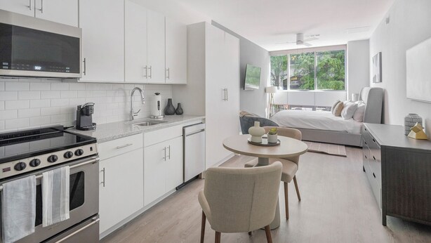 A modern kitchen with white cabinetry, a stove, and a sink. There is a dining area with a round table and chairs. The room has a large window with a view of greenery outside.
