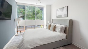A modern bedroom with a large bed, a TV, and a window with a view of trees.