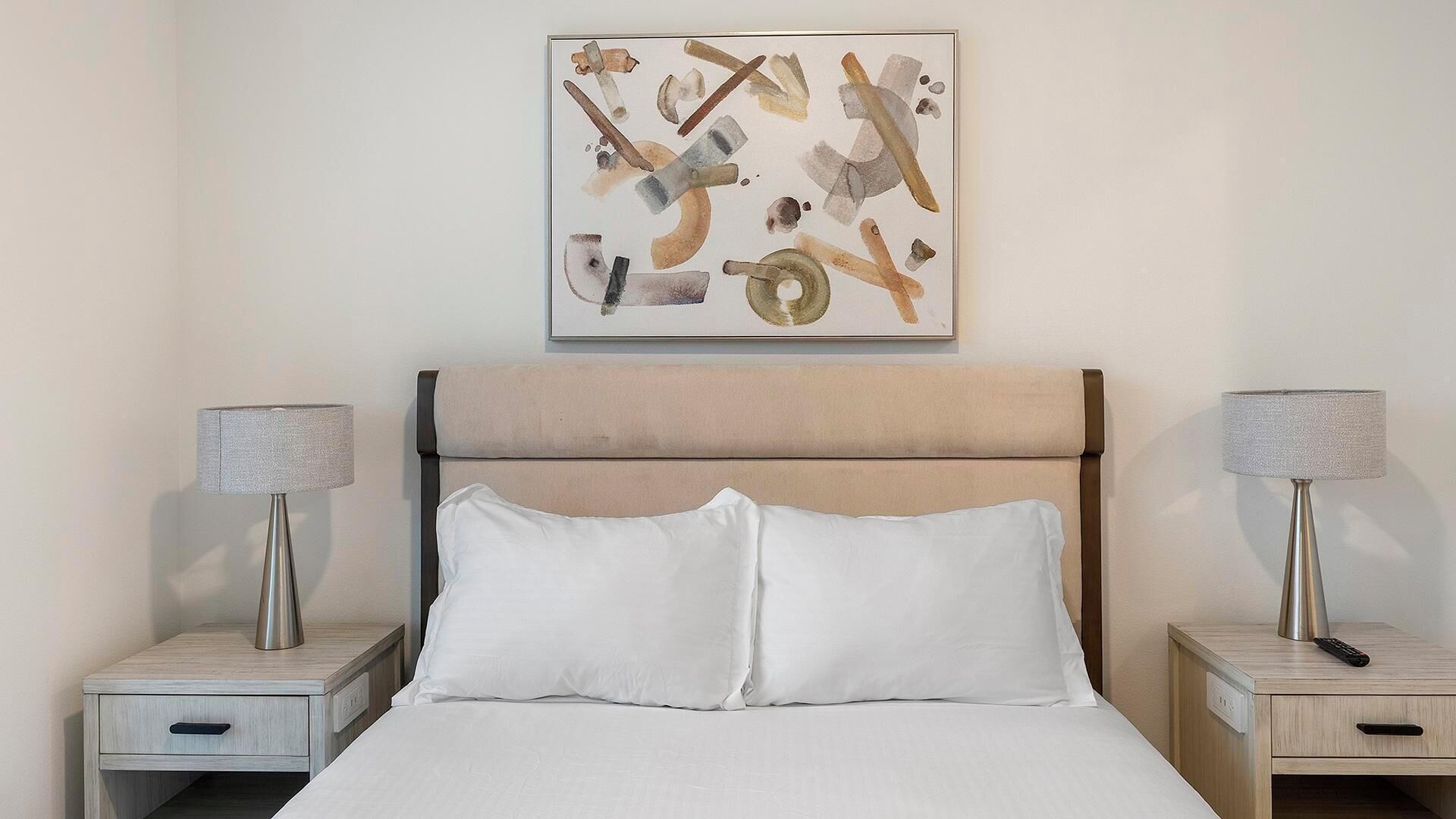 Une chambre d’hôtel avec un lit, deux tables de chevet avec des lampes et un tableau abstrait accroché au mur.
