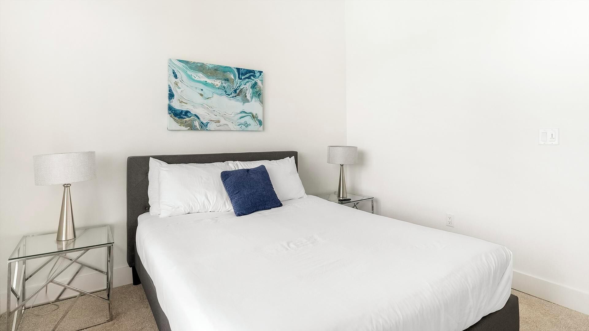 A neatly made bed with white bedding and a blue accent pillow, flanked by two bedside tables with lamps, in a minimalist room with a single abstract painting on the wall.