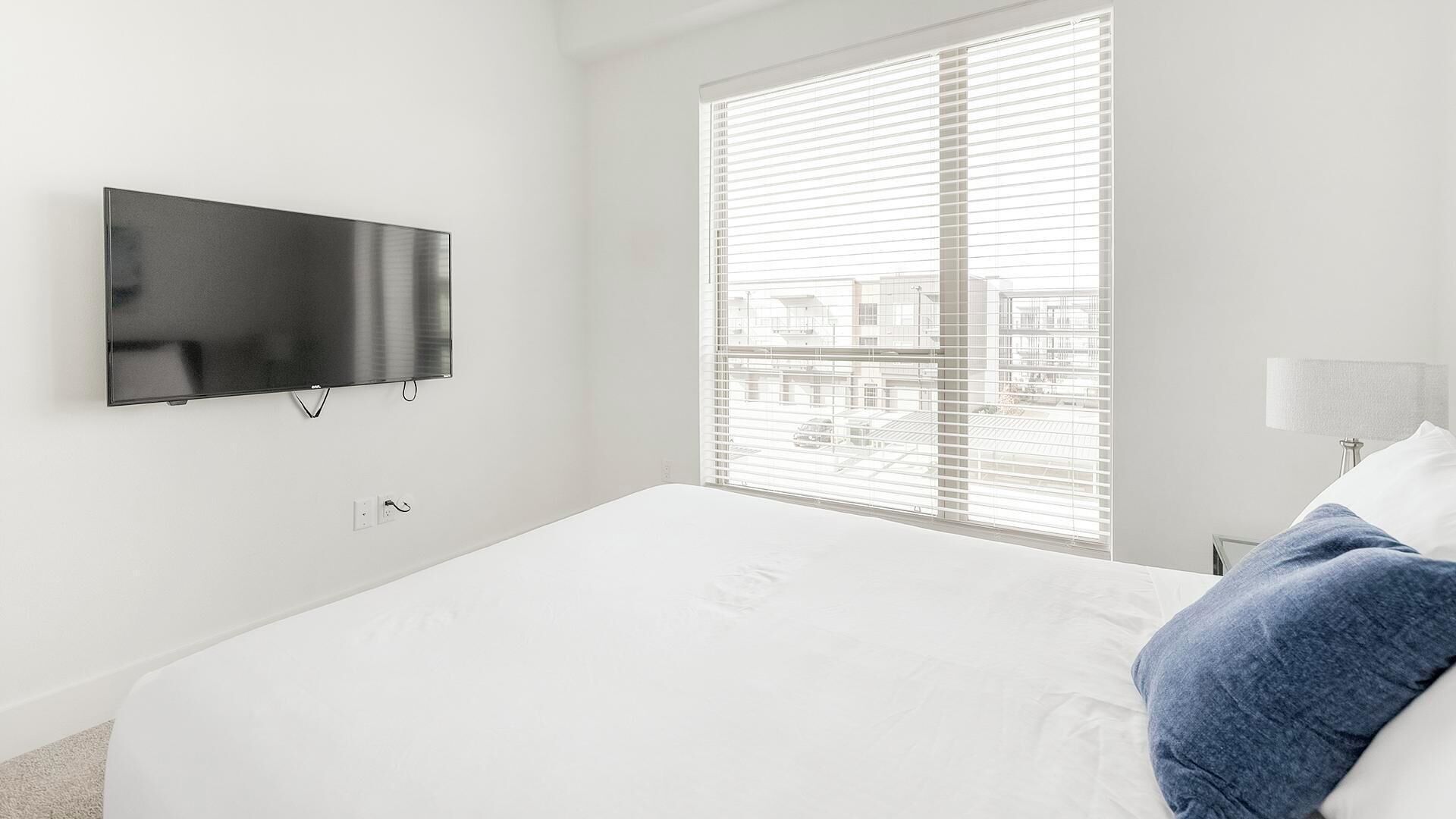 A modern hotel room with a large bed, a flat-screen TV mounted on the wall, and a window with blinds.